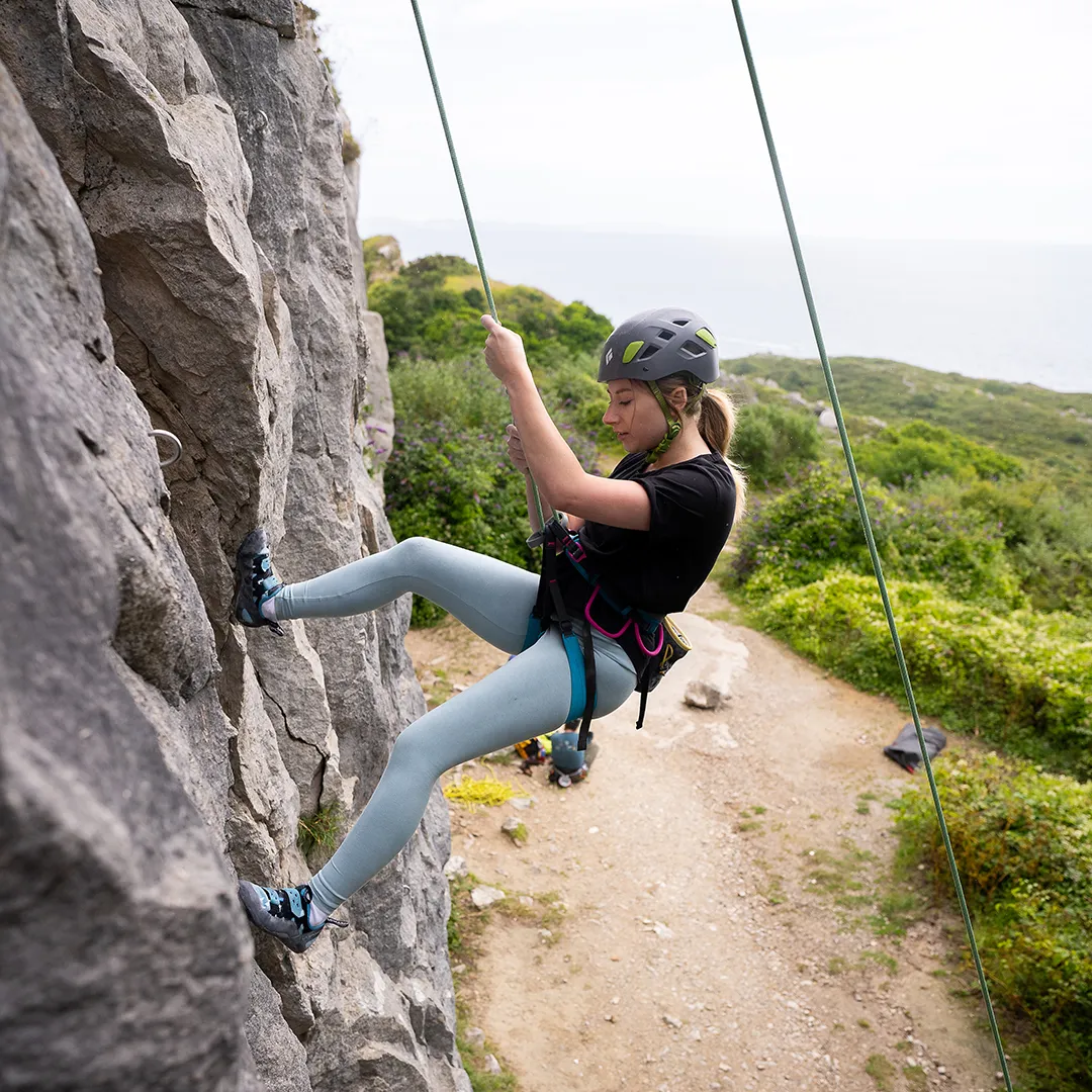 Laura is being lowered to the ground from the top of a wall. She has her legs extended with her feet out in front of her, as she walks down the wall.